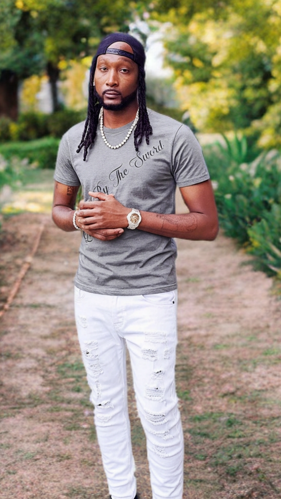 Man wearing a gray t-shirt and white pants standing outdoors with greenery in the background