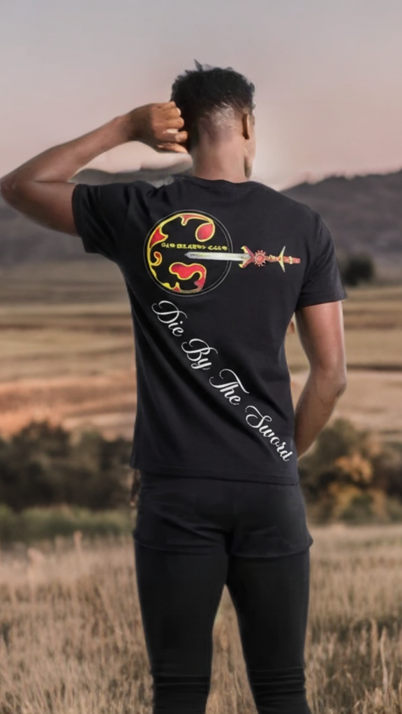 Person wearing a black t-shirt with a design and text, standing in a field with mountains in the background.